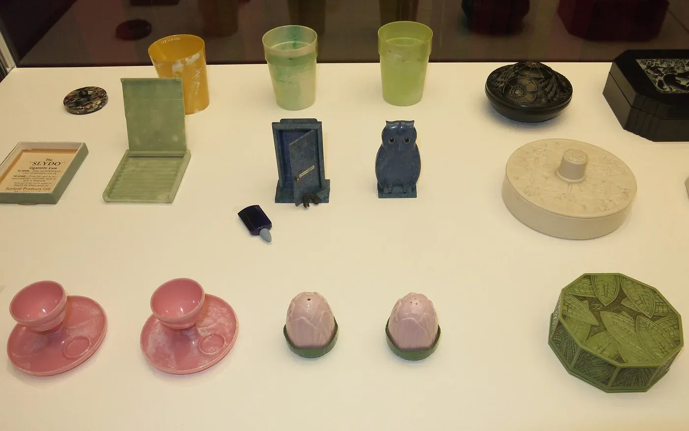A museum display case of small urea-formaldehyde household objects (cigarette cases, cups, an owl figurine, salt and pepper shakers, cup-and-saucer sets and trinket boxes) in yellow, green, blue, pink, cream and black.