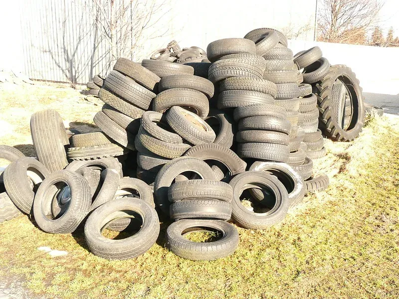 A large outdoor pile of worn car and tractor tires stacked against a corrugated metal fence, with bare trees in the background.