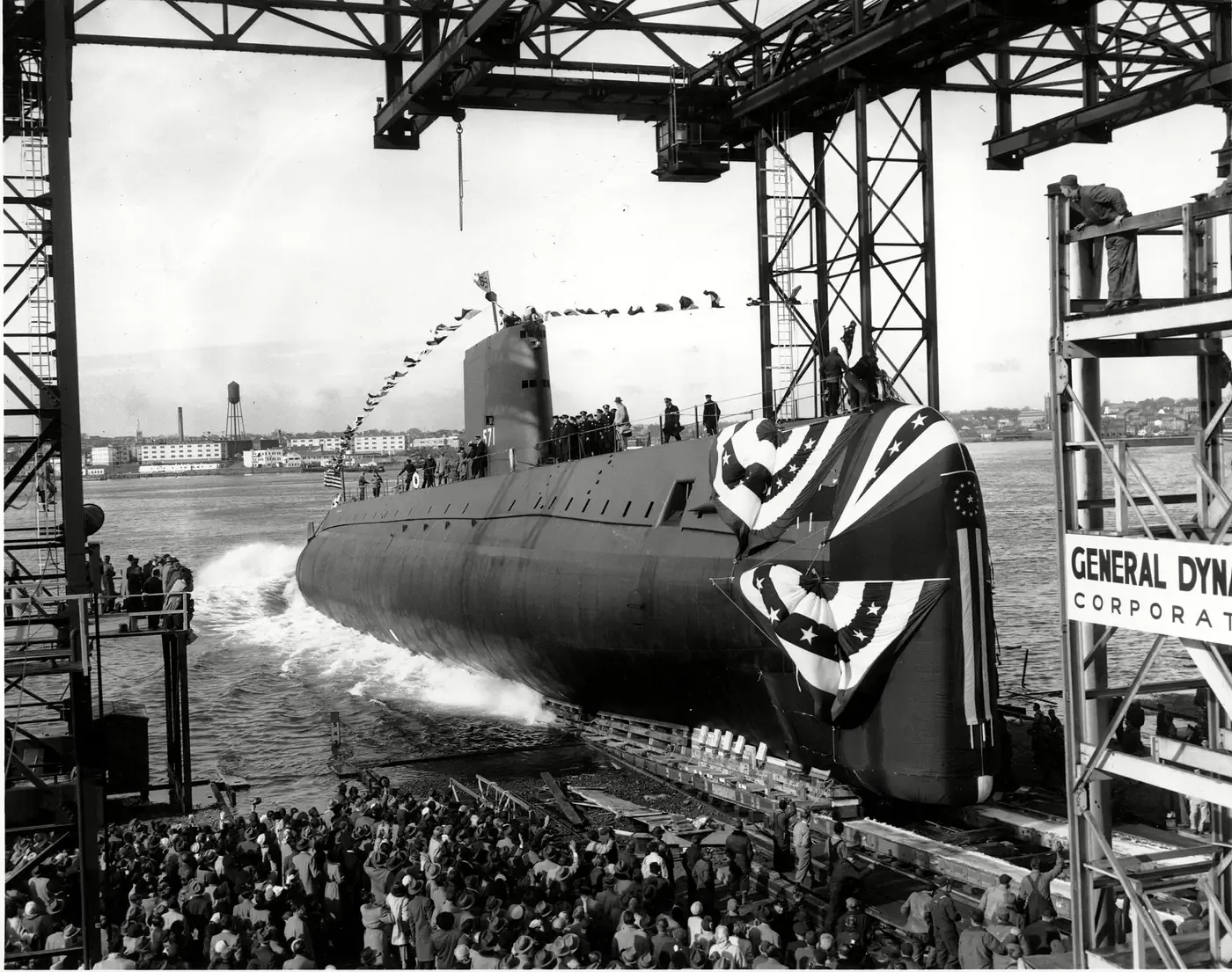 A submarine, decorated with bunting, sliding stern-first down a shipyard slipway into a river as a large crowd watches from the dock.