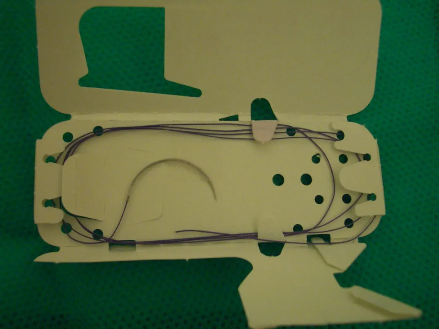 A packet of violet braided suture thread with a curved needle, coiled inside an opened foil and card package printed with perforated tear tabs.