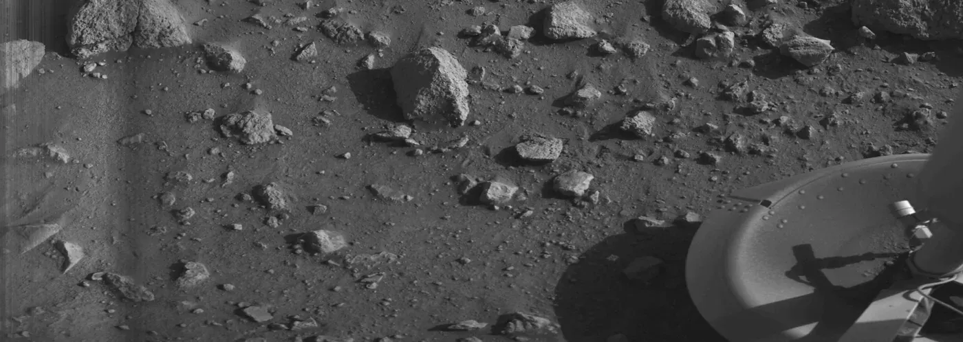 A grainy black-and-white photograph of a rock-strewn, sandy Martian plain, with the curved edge of a spacecraft footpad and its shadow visible in the lower right corner.