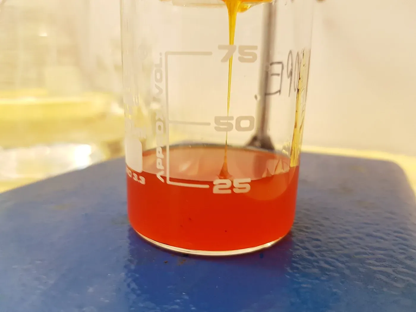 A glass beaker of thick, translucent orange-red liquid on a laboratory bench, with a stream of the same viscous liquid pouring into it from above.
