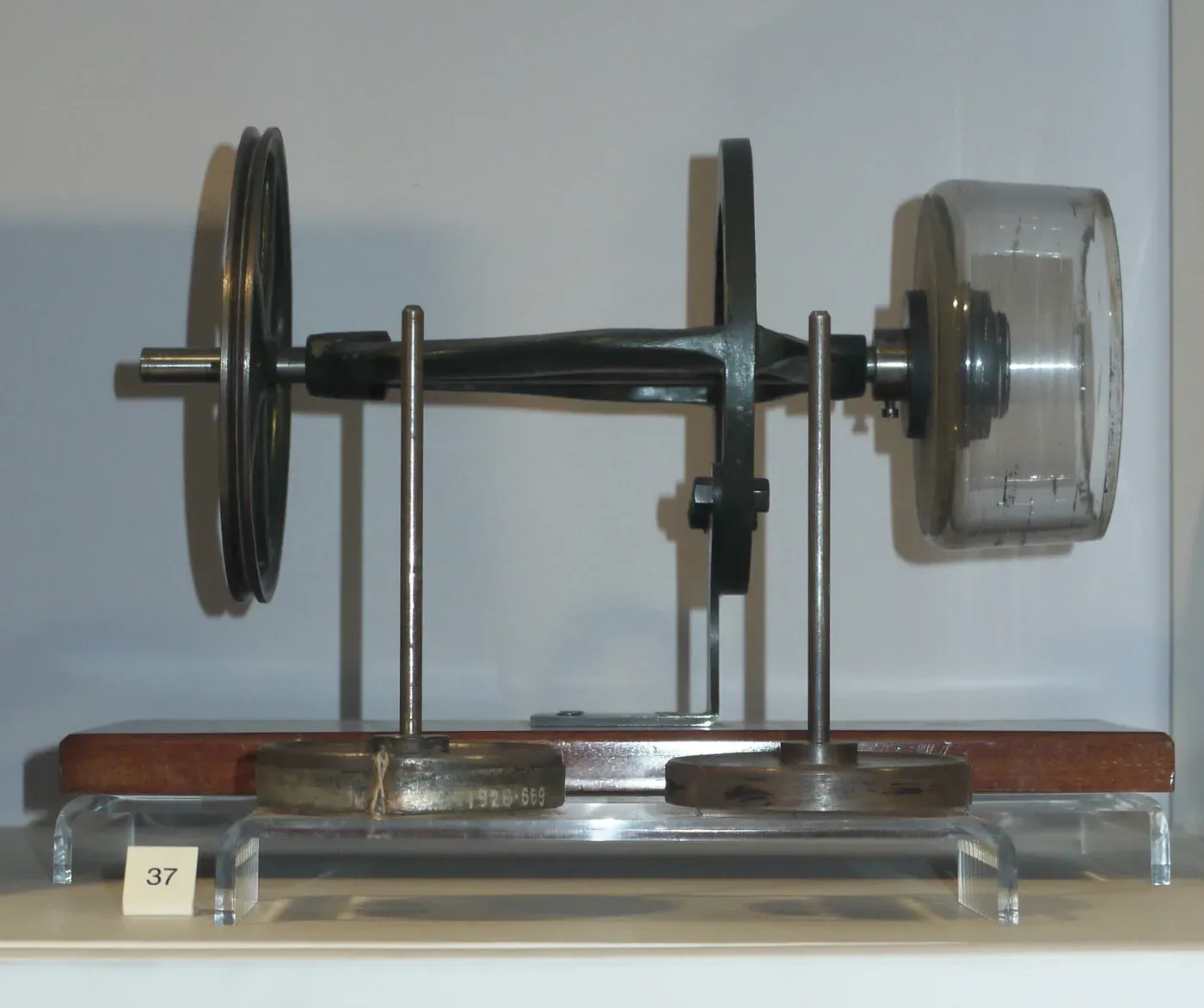 A small brass and glass demonstration spinning apparatus in a museum display case, with two large grooved pulleys, a drive shaft, and a glass bobbin mounted at one end.