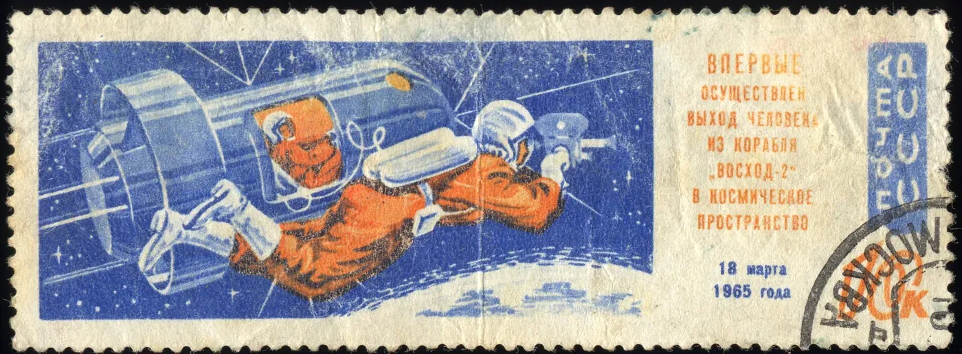 A 1965 Soviet postage stamp illustrating a cosmonaut in an orange spacesuit floating outside the cylindrical Voskhod 2 spacecraft against a starry blue background, with Cyrillic text and the date 18 March 1965.