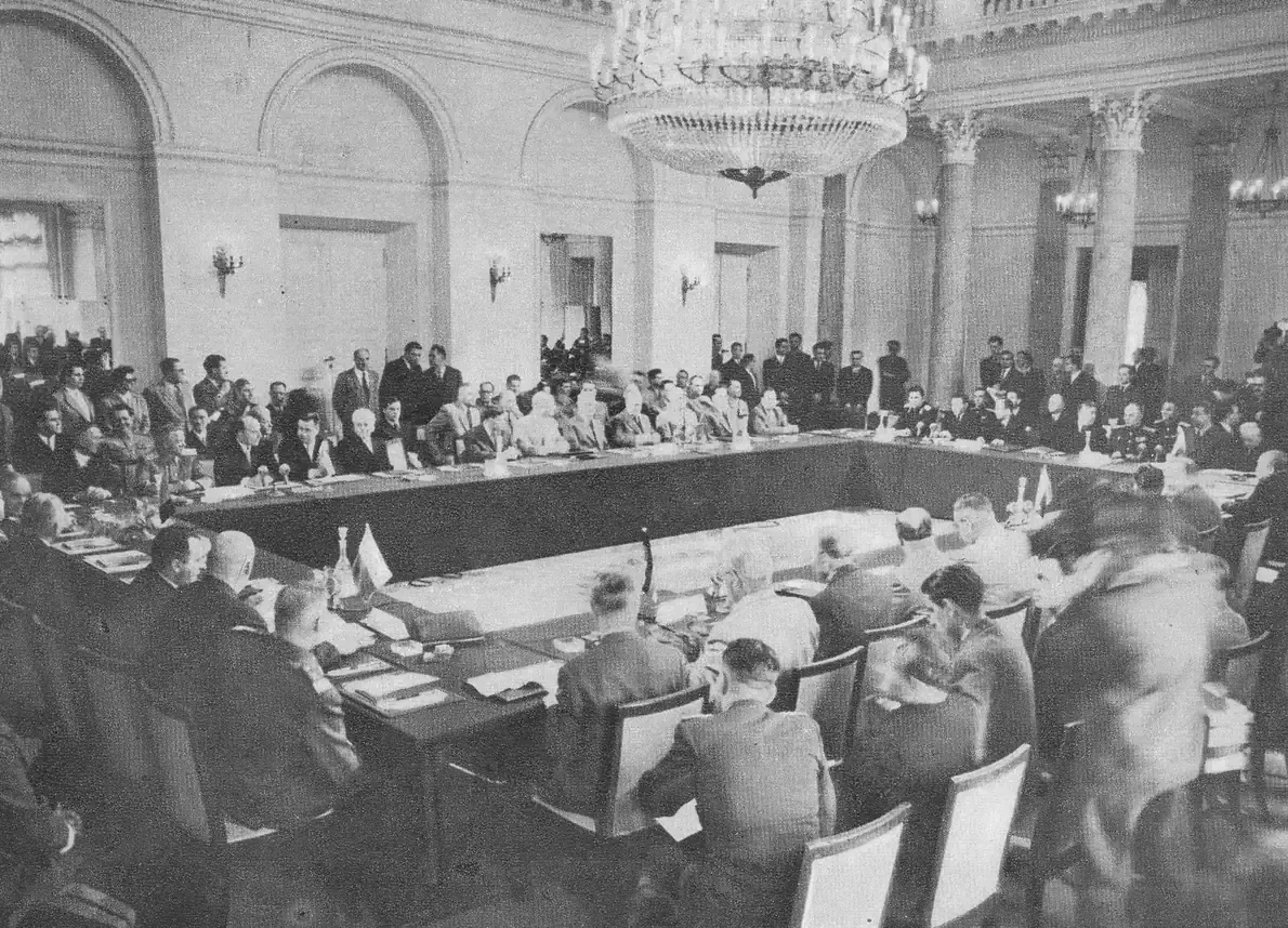 A large formal conference hall with a long U-shaped table of seated delegates in suits and military uniforms, beneath ornate chandeliers and classical columns.