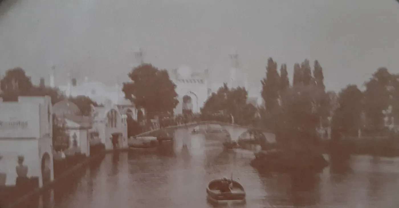 A sepia-toned view across an ornamental lake at an exhibition ground, with white pavilions, a footbridge and small boats on the water.