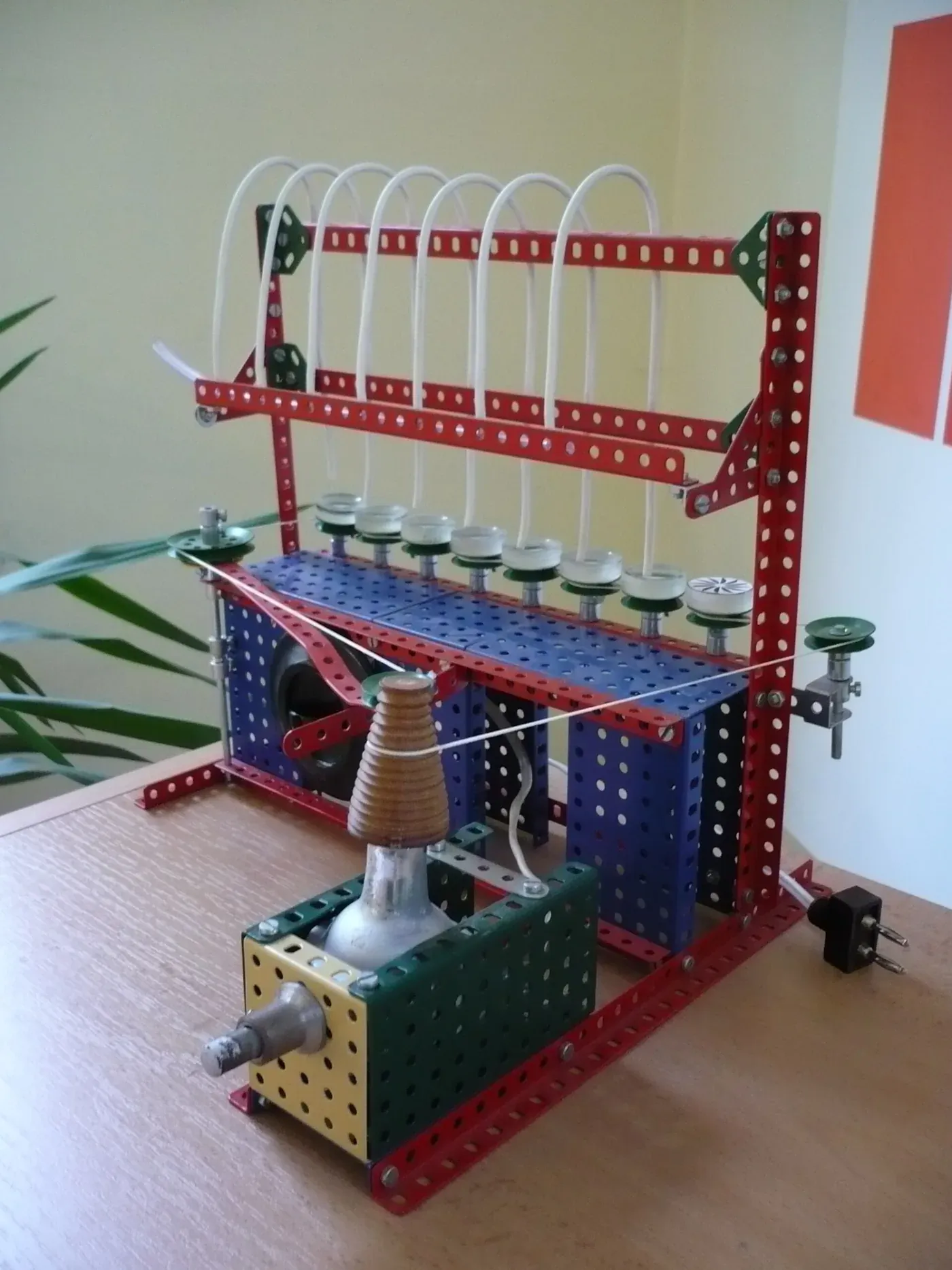 A tabletop machine built entirely from red, blue, green and yellow perforated toy-construction-kit strips, with a small motor, a row of small moulds along one crossbar, and thin tubes feeding down to them.