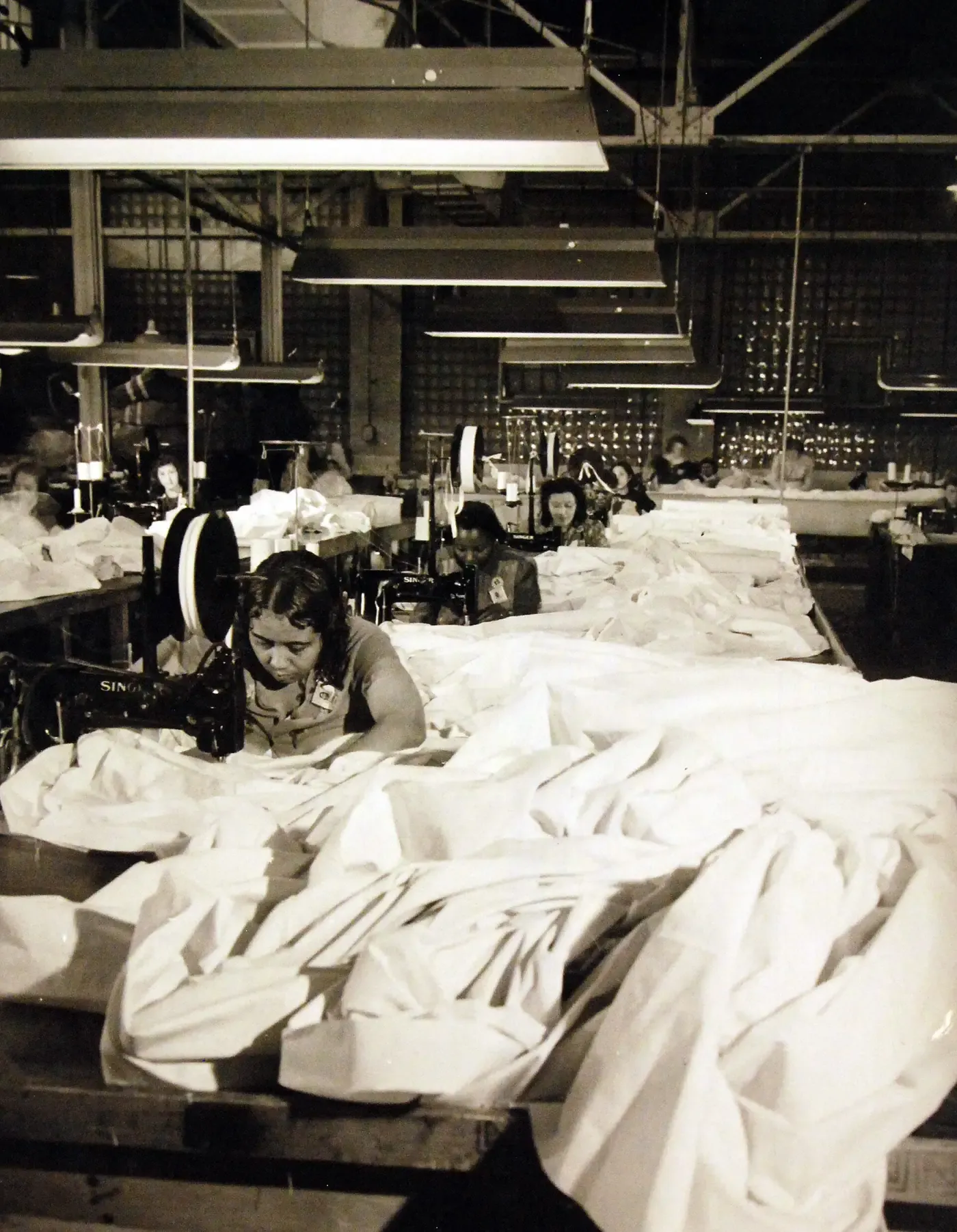 A large industrial sewing room with rows of workers at Singer sewing machines, working with billowing white fabric that covers most of the tables in the foreground, under long fluorescent light fixtures.