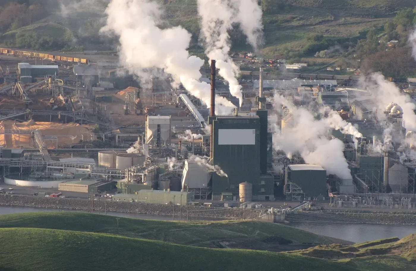 An aerial view of a large industrial wood pulp mill beside a river, with tall smokestacks releasing plumes of white steam over rows of green processing buildings and stacked timber.