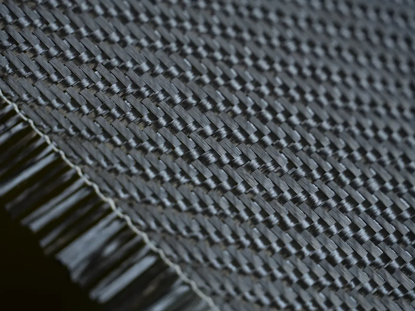 A close-up of tightly woven black carbon fiber fabric, its diagonal twill pattern catching the light, with loose frayed strands along one cut edge.