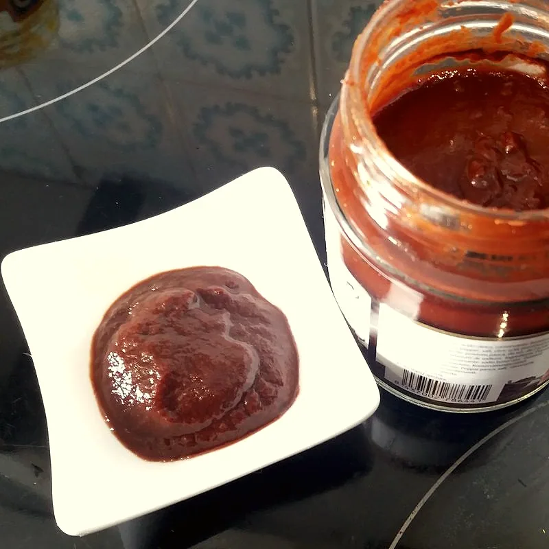 A dollop of dark red ají panca paste on a small dish beside its open jar