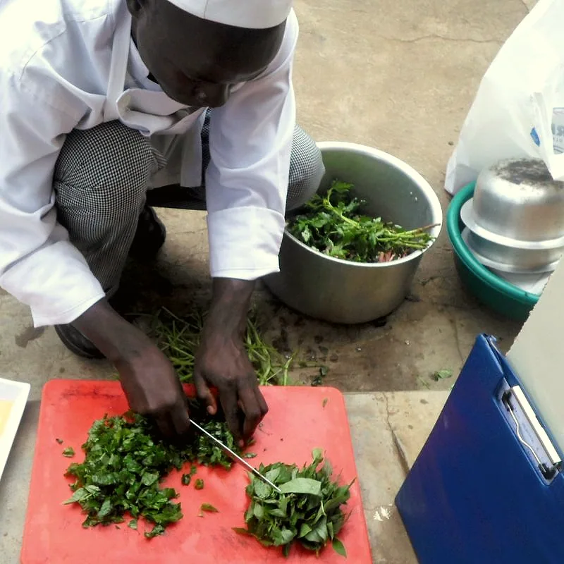 A cook chopping fresh green amaranth leaves on a red cutting board, with a metal bowl of whole leaves nearby