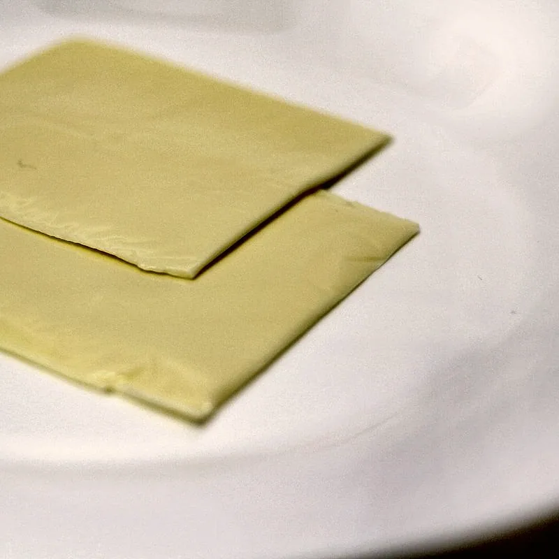 Two square unwrapped slices of pale yellow American cheese on a white plate