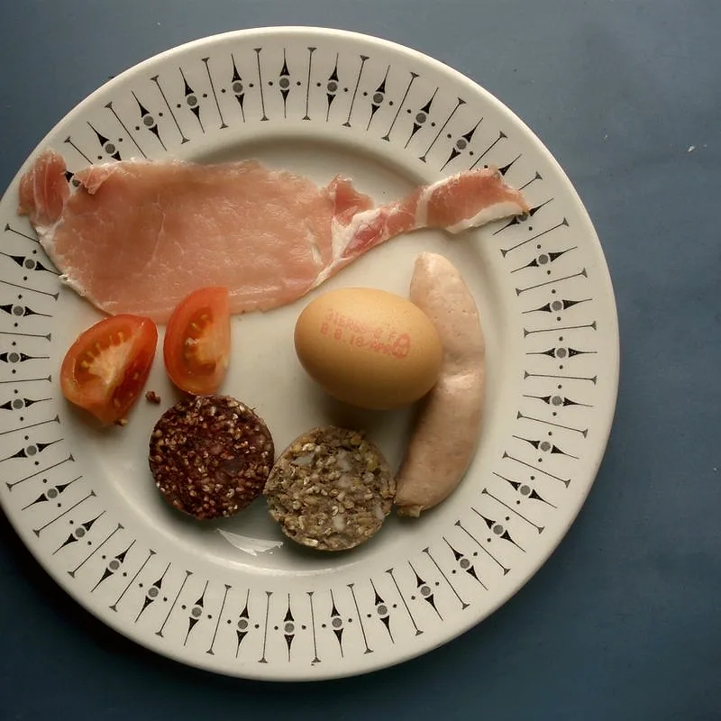 A raw back bacon rasher, a round eye of pink meat with a streaky fat tail, on a plate alongside other raw Irish breakfast ingredients