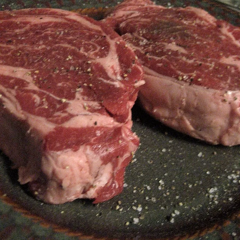 Two raw, well-marbled beef chuck steaks seasoned with salt and pepper on a dark plate