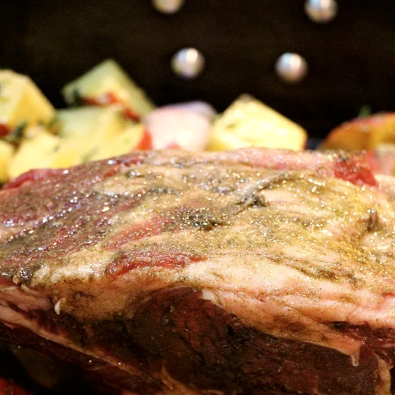 A raw, seasoned bone-in beef rib roast, resting over a bed of potatoes and shallots