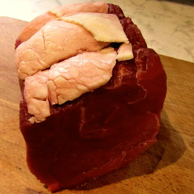 A raw, lean beef topside roasting joint with a thin cap of fat, on a wooden board
