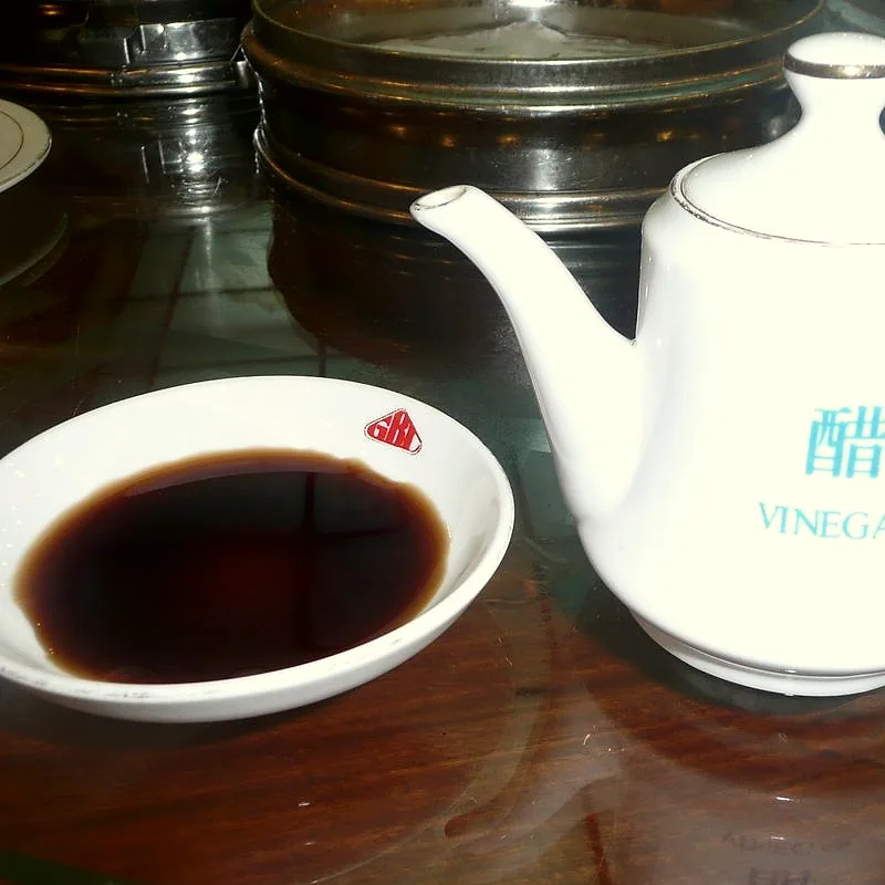 A small dish of dark Chinese black vinegar beside a white teapot-style vinegar pot labelled 醋 VINEGAR
