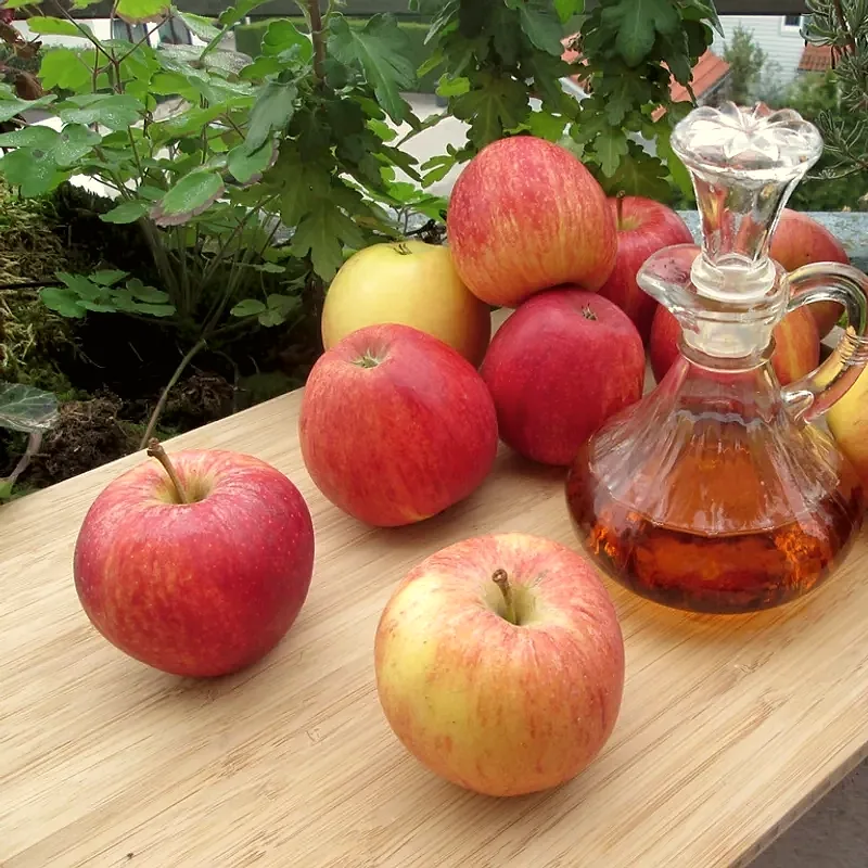 A glass decanter of amber apple cider vinegar beside several whole red and yellow apples on a wooden table