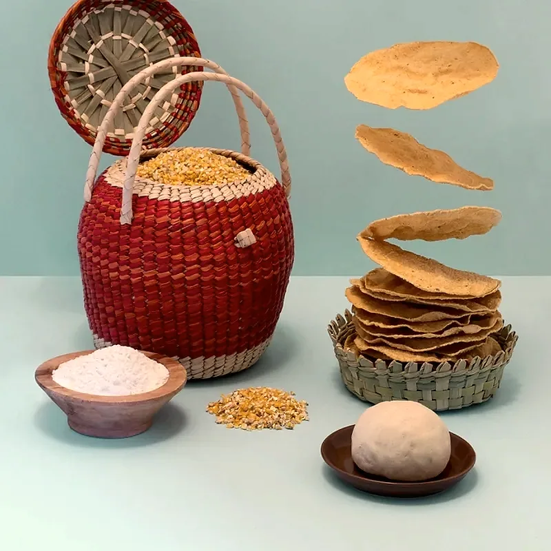Stacked corn tortillas in a woven basket, with fresh masa dough, ground corn and corn kernels beside a basket of dried corn