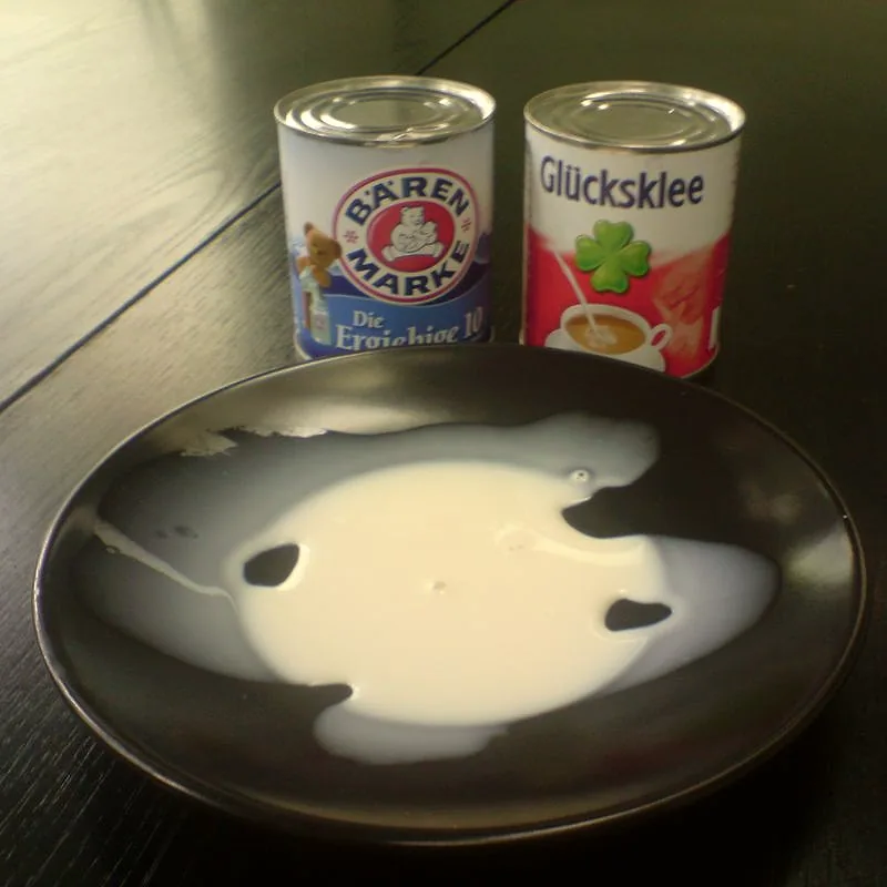 A pool of thick white evaporated milk poured onto a dark plate, with two tins of evaporated milk in the background