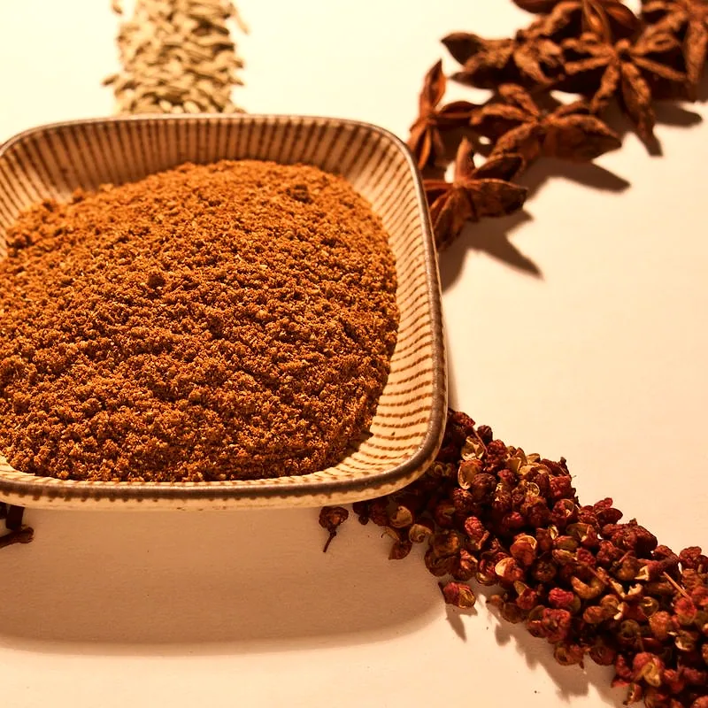 A dish of reddish-brown five spice powder surrounded by its whole spice ingredients including star anise and Sichuan peppercorns
