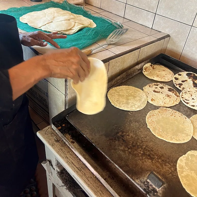 Flour tortillas cooking on a hot griddle, with a raw dough ball being pressed