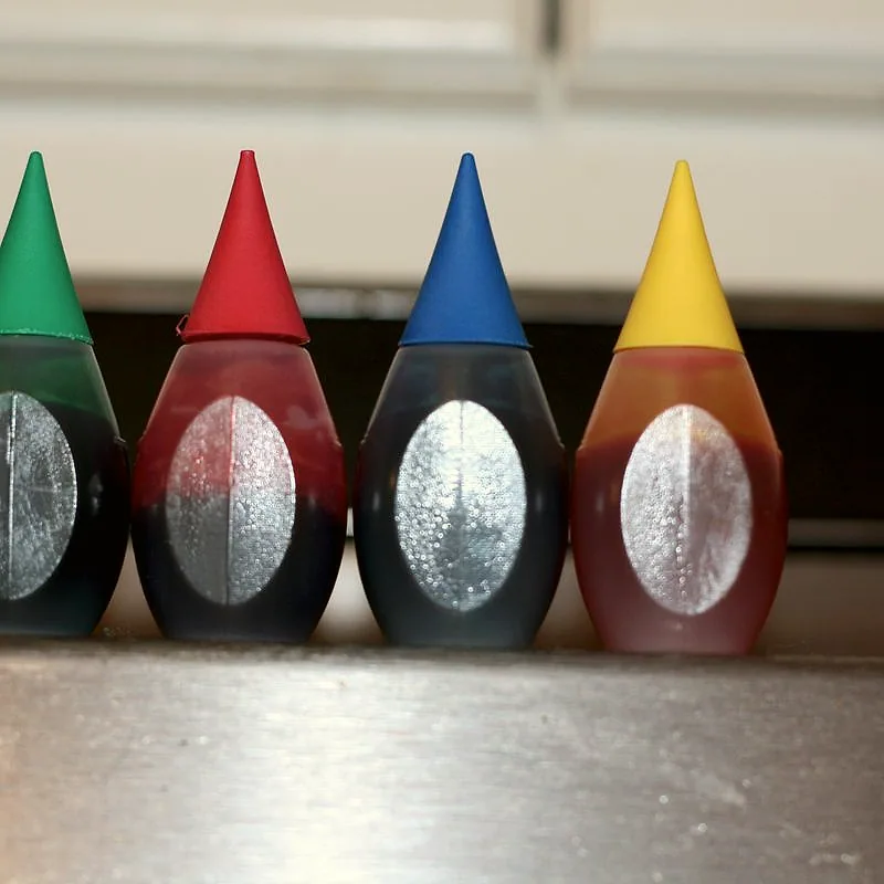 Four small bottles of liquid food colouring in green, red, blue, and yellow, lined up with their colour-coded caps