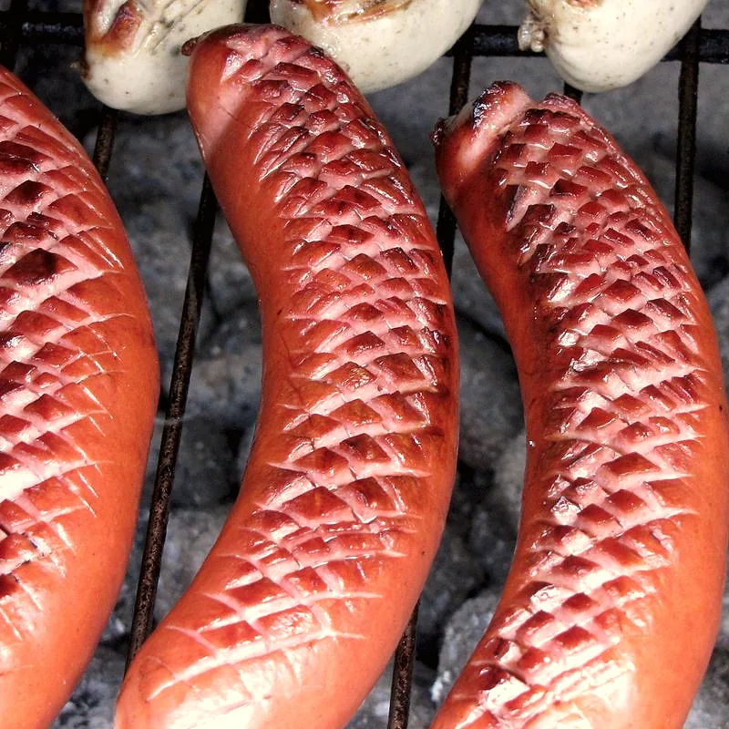 Scored frankfurter sausages grilling on a barbecue rack over charcoal