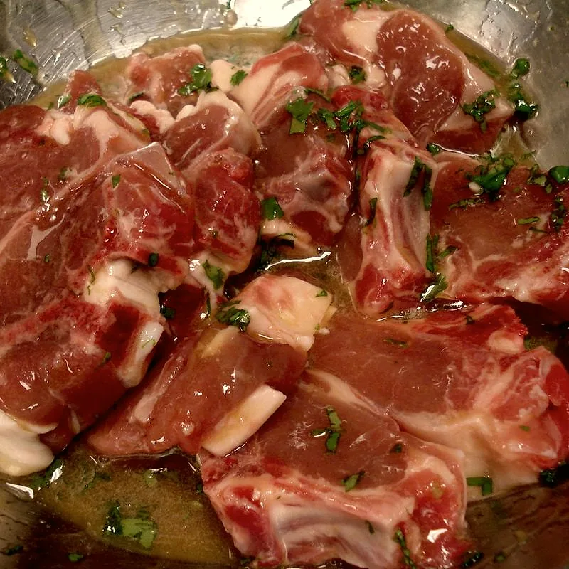 Raw goat chops marinating in oil with chopped herbs in a metal bowl