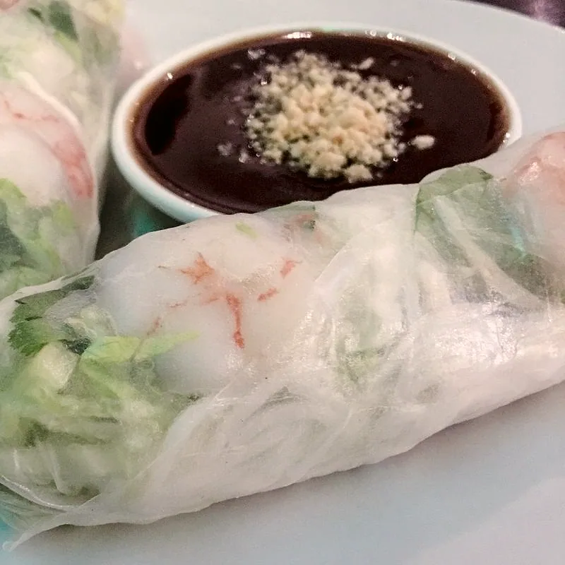 A small bowl of dark brown hoisin sauce topped with crushed peanuts, beside a Vietnamese spring roll
