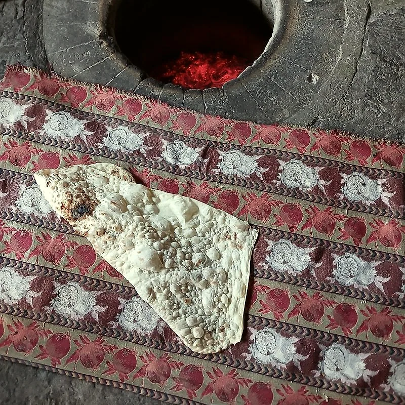 A thin, bubbled sheet of lavash bread resting beside a traditional tonir oven pit