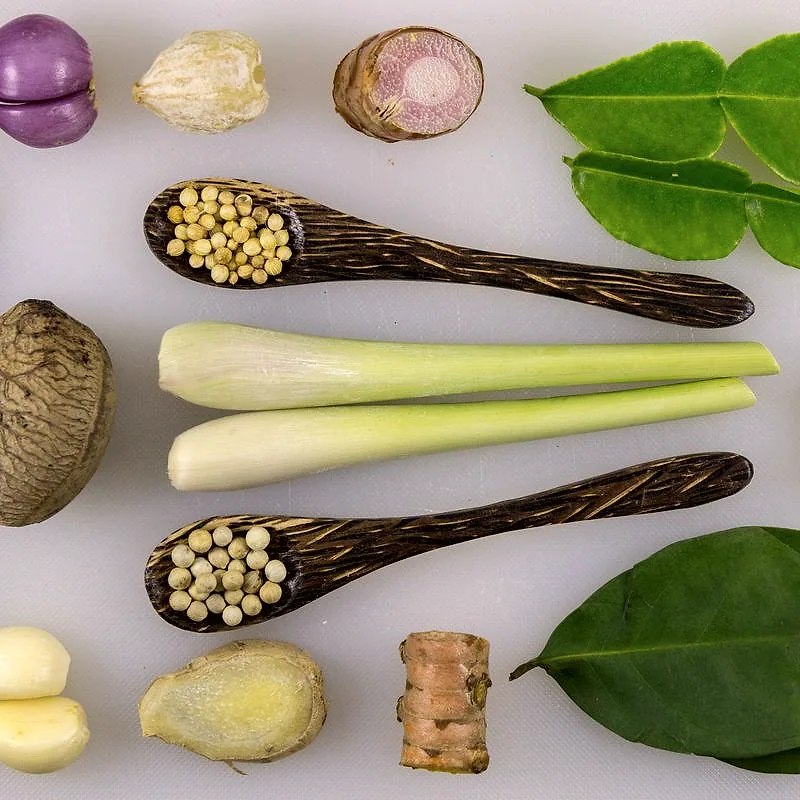 Two trimmed pale lemongrass stalks laid across wooden spoons, alongside other spices on a chopping board