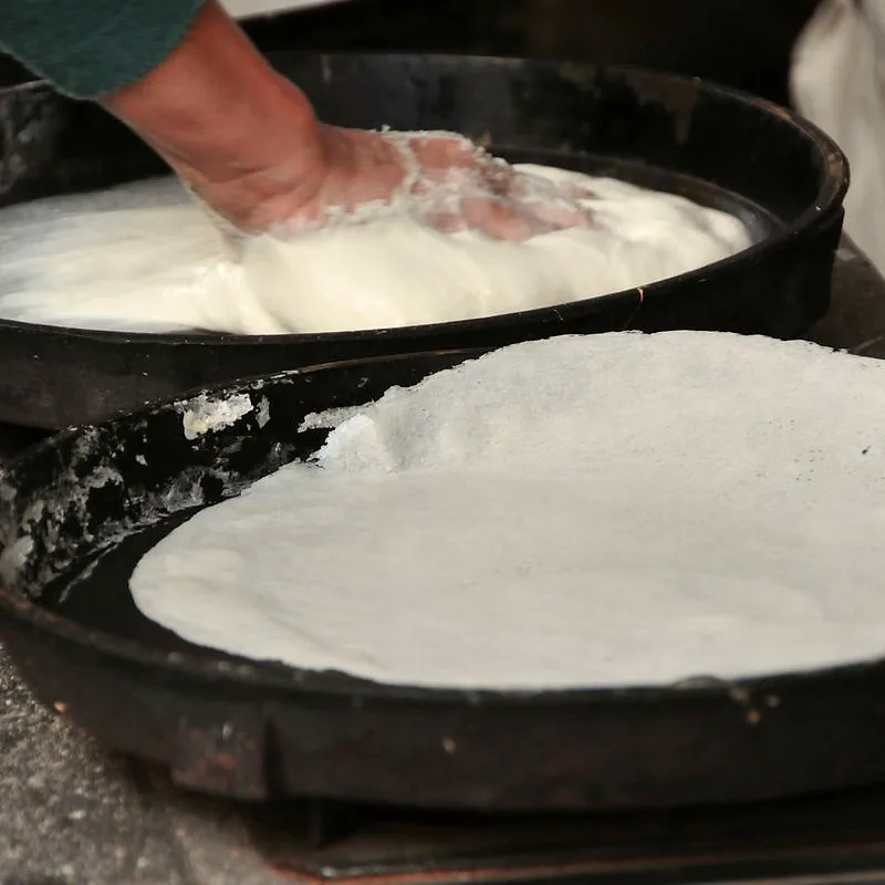 A thin, fresh wheat spring-roll wrapper cooking on a hot flat pan, with a hand smoothing more batter into a wrapper on a second pan