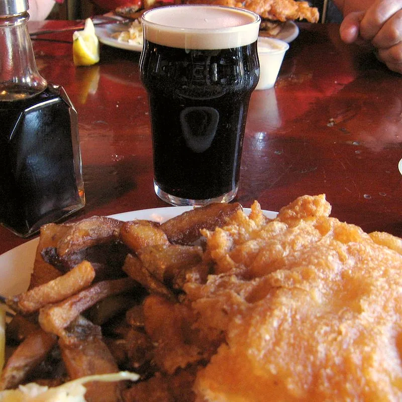 A glass bottle of dark malt vinegar on a table beside a plate of fish and chips