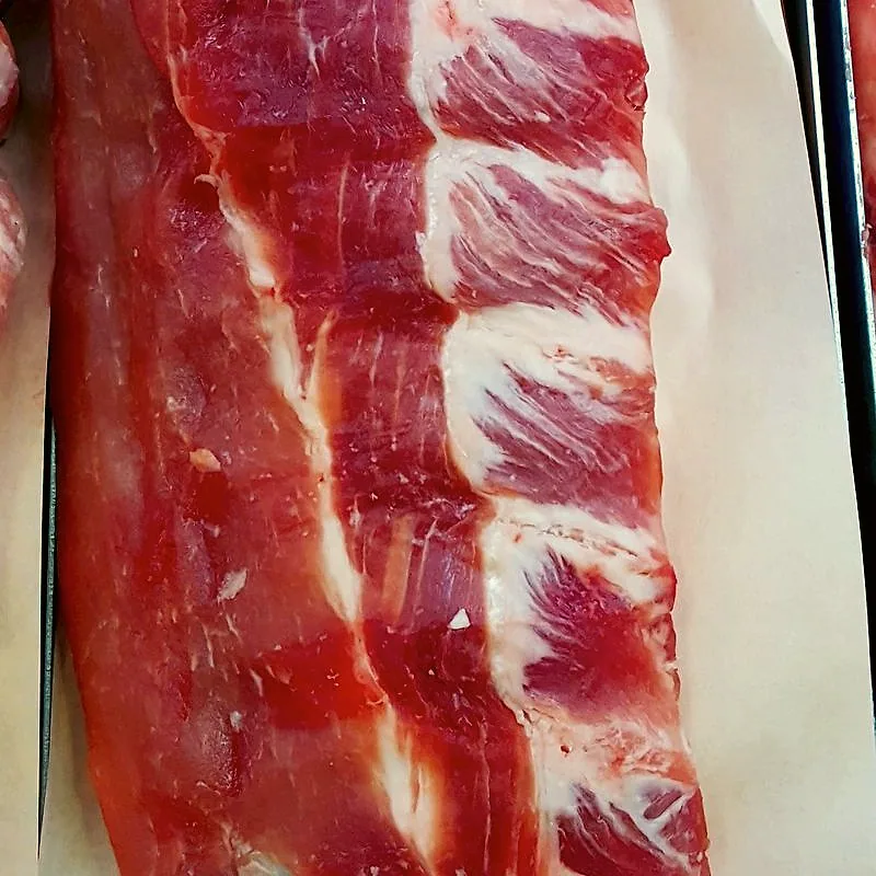 A raw rack of pork baby back ribs, meat-side up, showing dark red muscle between the rib bones on butcher paper