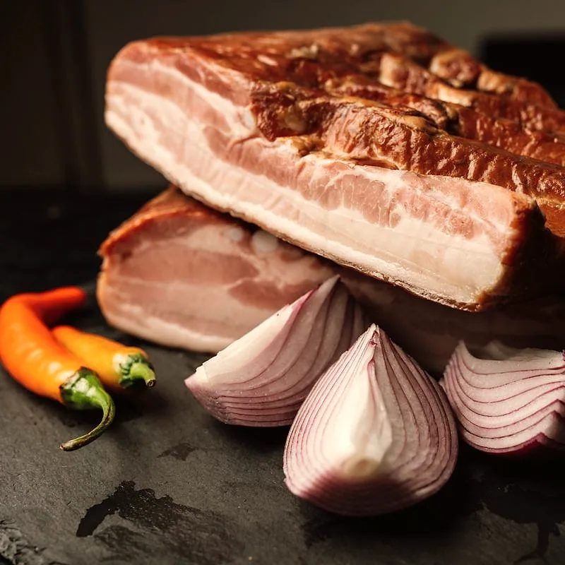 A raw slab of pork belly with distinct layers of fat and lean meat, beside sliced red onion and chillies