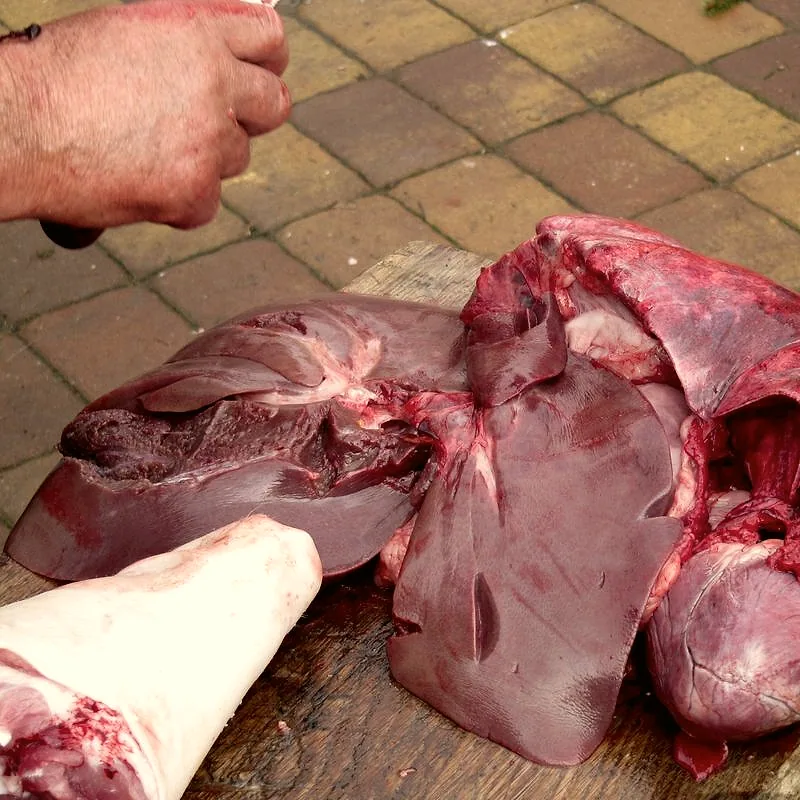 A raw, dark reddish-brown pig's liver laid on a wooden board, freshly butchered alongside other offal