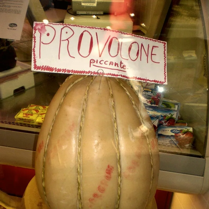 A whole pale golden provolone cheese, bound with string in its traditional rounded shape, on display in a deli case with a handwritten label