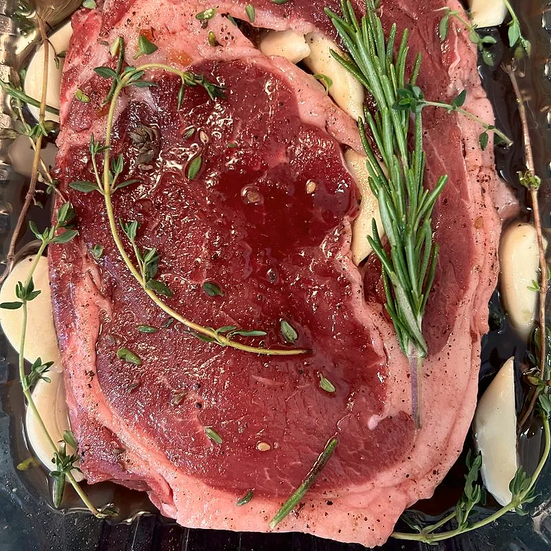 A raw ribeye steak marinating with garlic cloves, rosemary and thyme sprigs