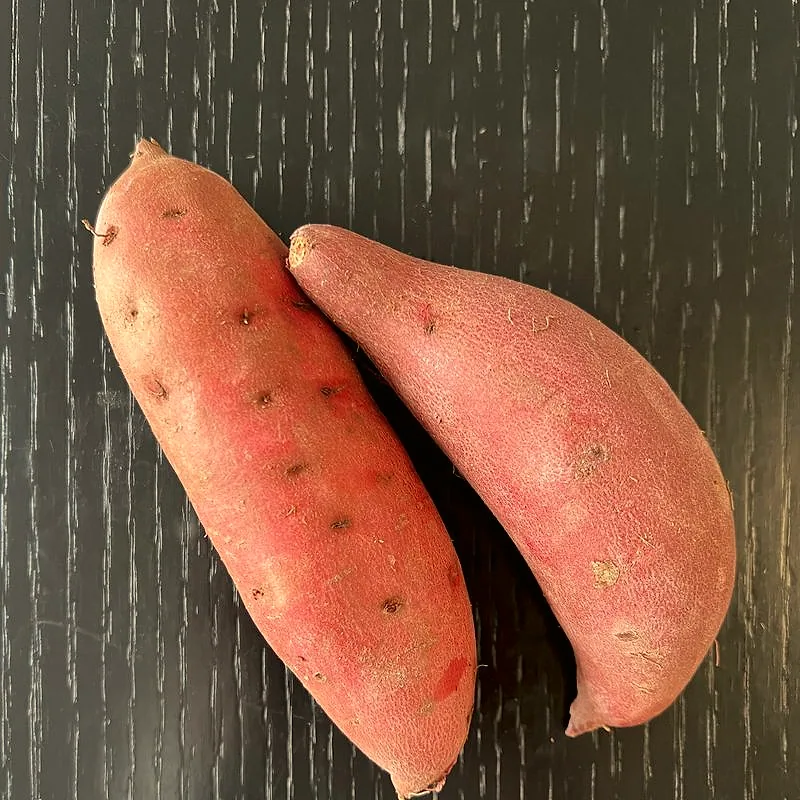 Two whole raw sweet potatoes with pink-orange skin on a dark wooden surface