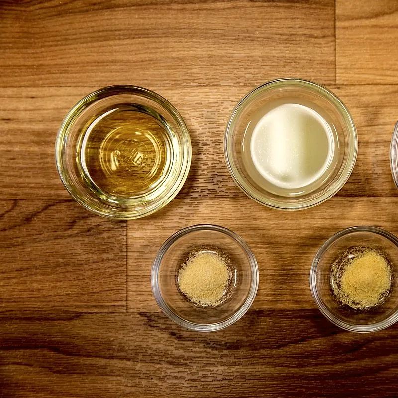A small glass bowl of pale golden vegetable oil, viewed from above beside bowls of other ingredients on a wooden surface