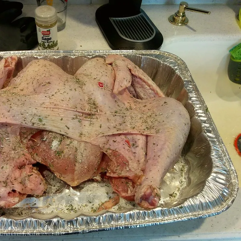 A raw whole turkey seasoned with dried herbs, sitting in a foil roasting tray before cooking
