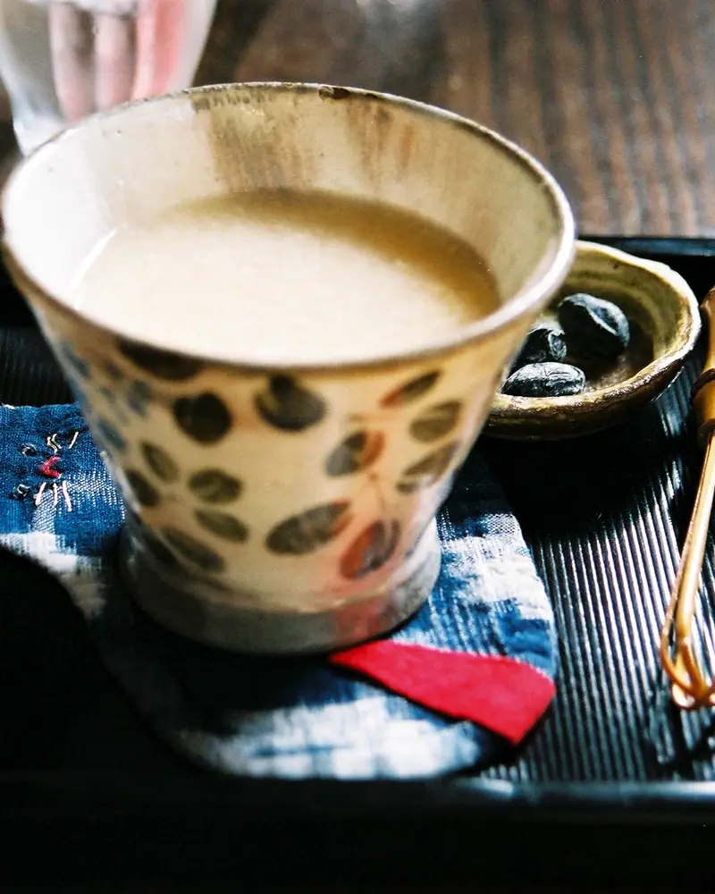 Pale beige amazake served warm in a hand-painted ceramic cup on a tray, with a small dish and red accent cloth beside it