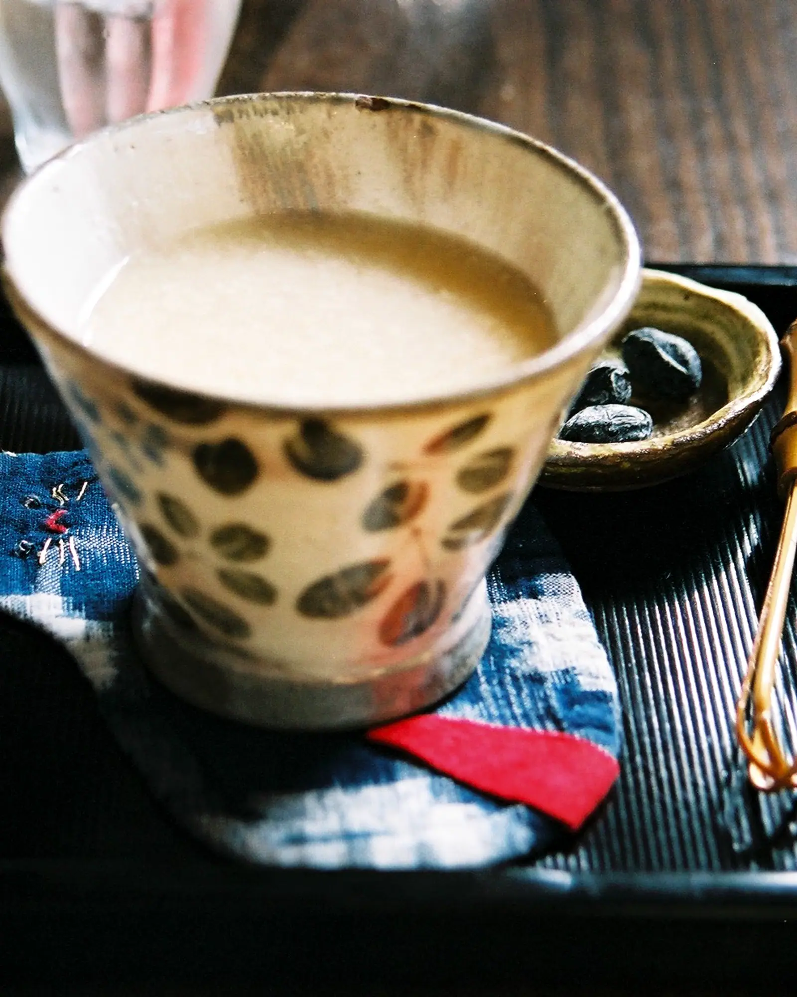Pale beige amazake served warm in a hand-painted ceramic cup on a tray, with a small dish and red accent cloth beside it
