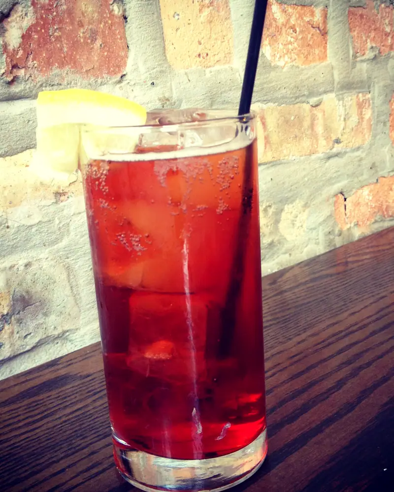 A red Americano cocktail in a tall glass over ice, garnished with a lemon wedge, on a dark wood bar