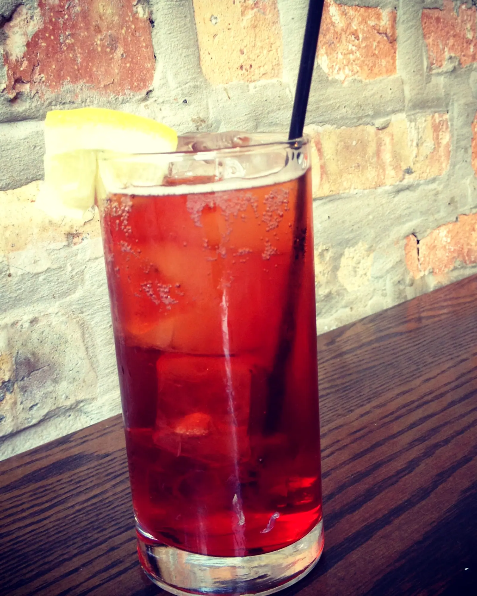 A red Americano cocktail in a tall glass over ice, garnished with a lemon wedge, on a dark wood bar
