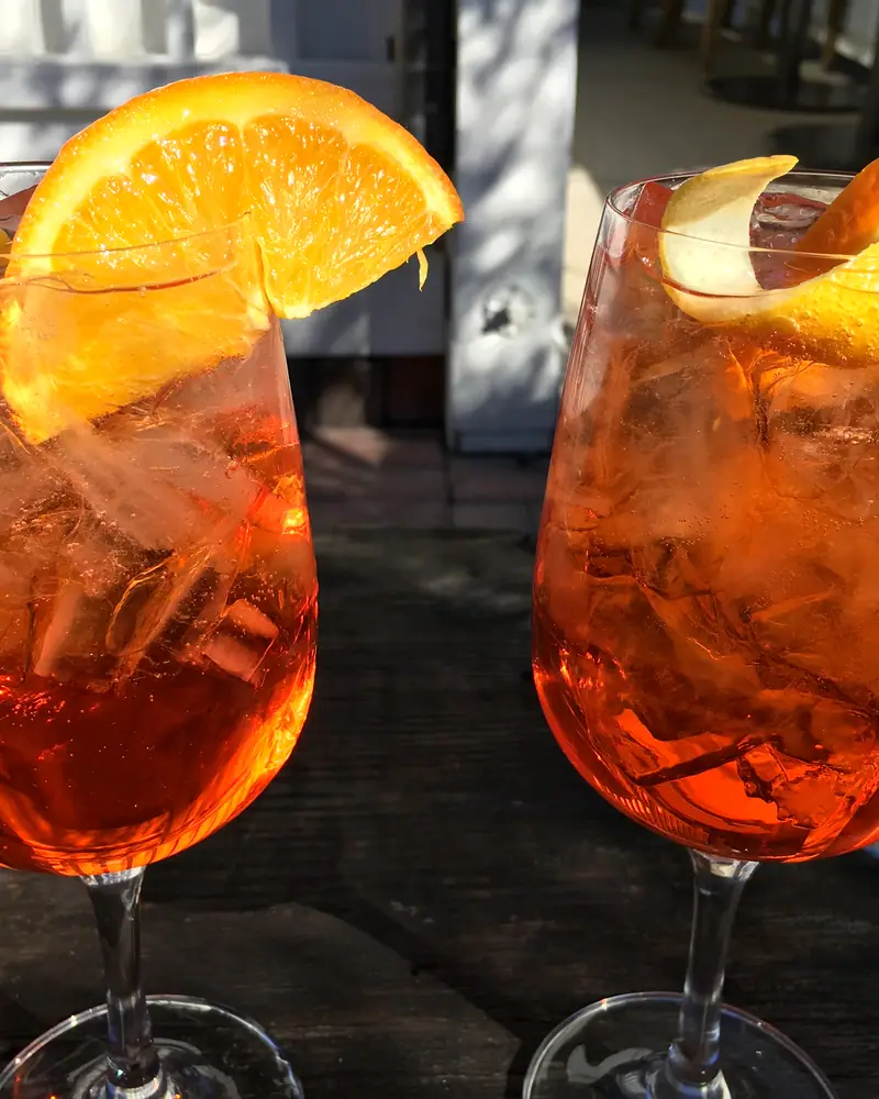 Two Aperol Spritz cocktails in large wine glasses over ice, garnished with orange slices