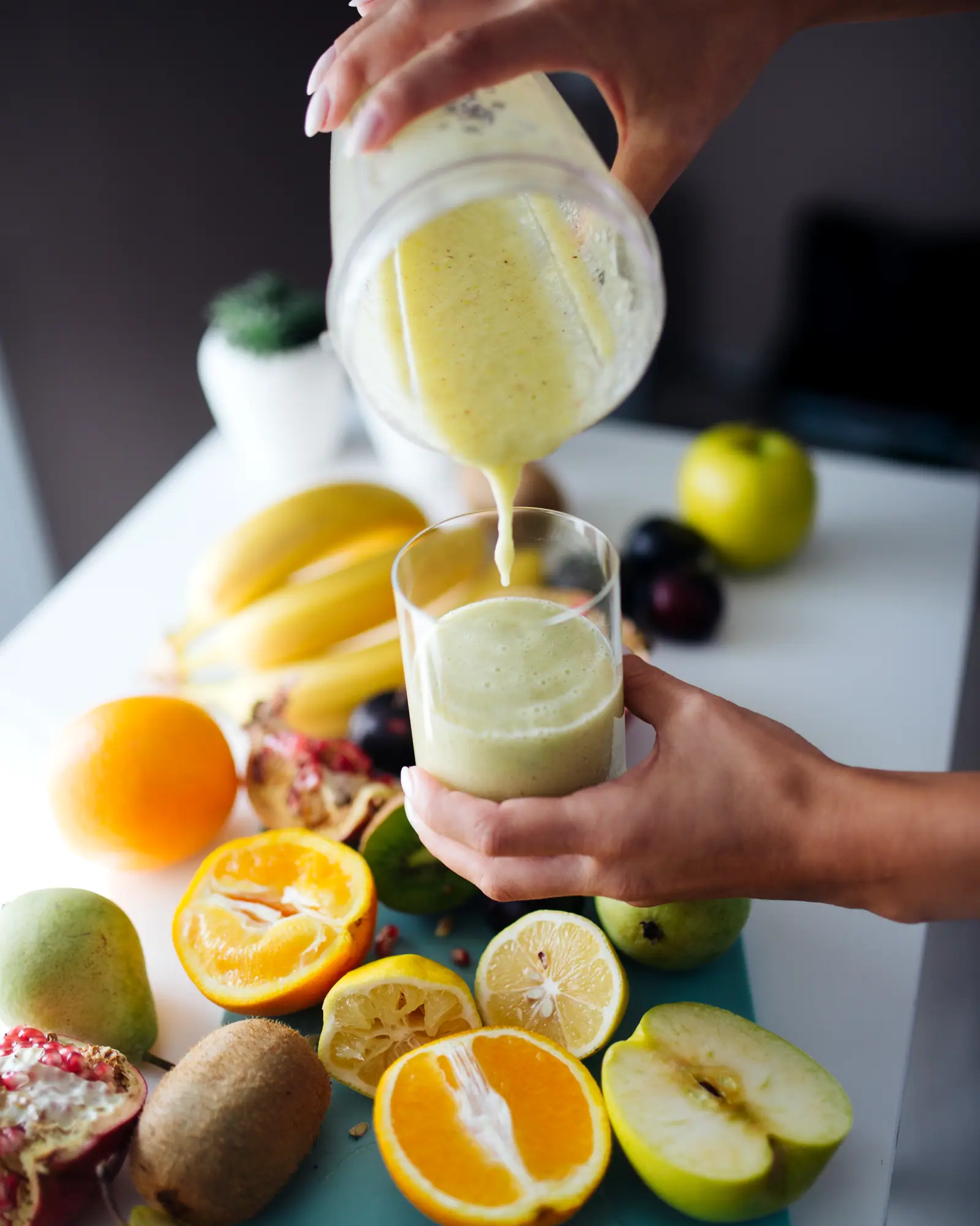 A pale yellow fruit smoothie being poured into a glass, surrounded by fresh fruit