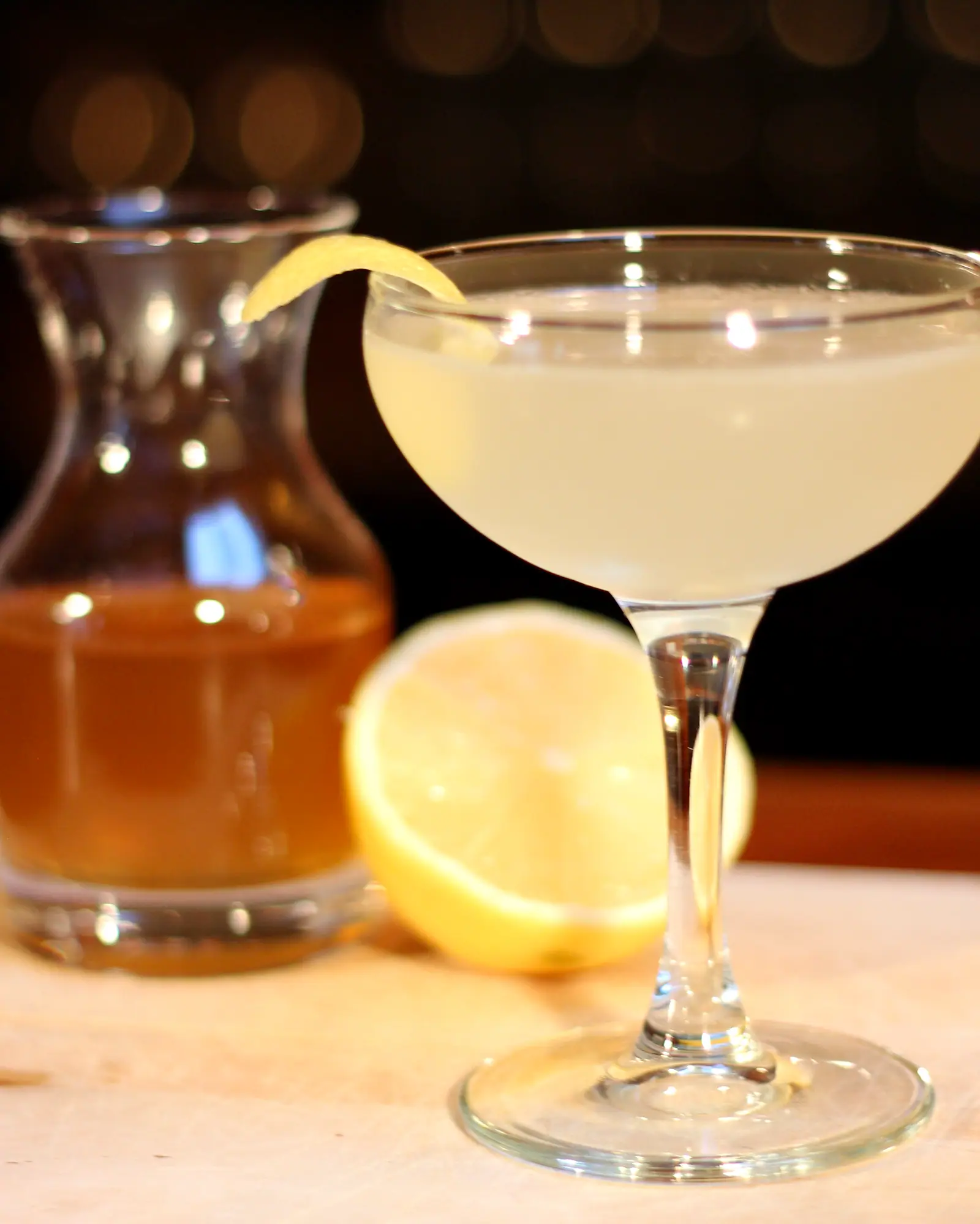 A Bee's Knees cocktail in a coupe glass with a lemon twist, pale golden-yellow, with lemon halves and a carafe of amber liquid in the background